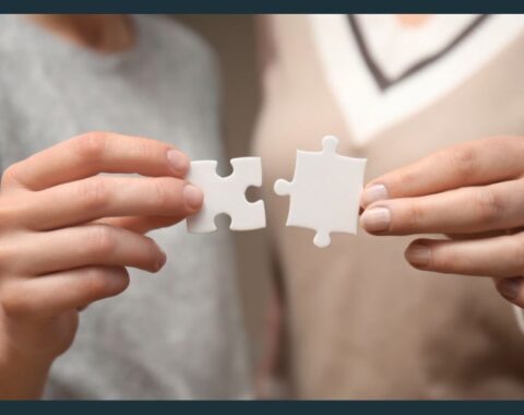 A photograph showcases two individuals engaged in a thoughtful conversation while holding interlocking puzzle pieces. The image symbolises collaboration, problem-solving, and a human-centred approach. The setting is clean and professional, with warm lighting that adds a sense of connection and empathy.