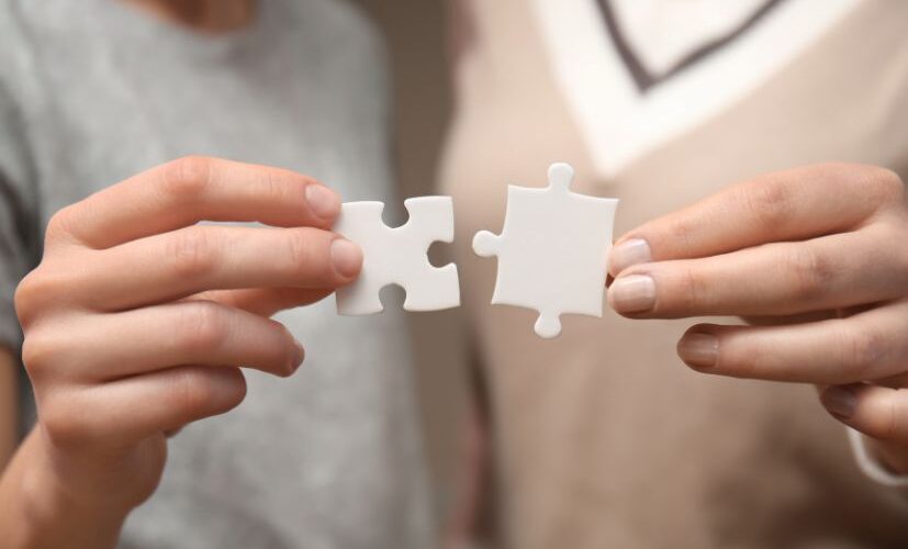 A photograph showcases two individuals engaged in a thoughtful conversation while holding interlocking puzzle pieces. The image symbolises collaboration, problem-solving, and a human-centred approach. The setting is clean and professional, with warm lighting that adds a sense of connection and empathy.