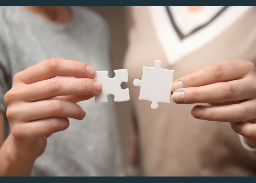 A photograph showcases two individuals engaged in a thoughtful conversation while holding interlocking puzzle pieces. The image symbolises collaboration, problem-solving, and a human-centred approach. The setting is clean and professional, with warm lighting that adds a sense of connection and empathy.
